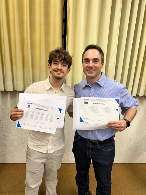 Duas pessoas exibindo certificados de reconhecimento em ambiente acadêmico.